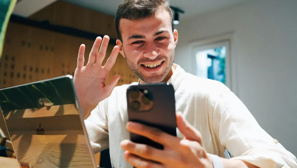 A smiling man waving at his smartphone while on a video call at a desk with a laptop.